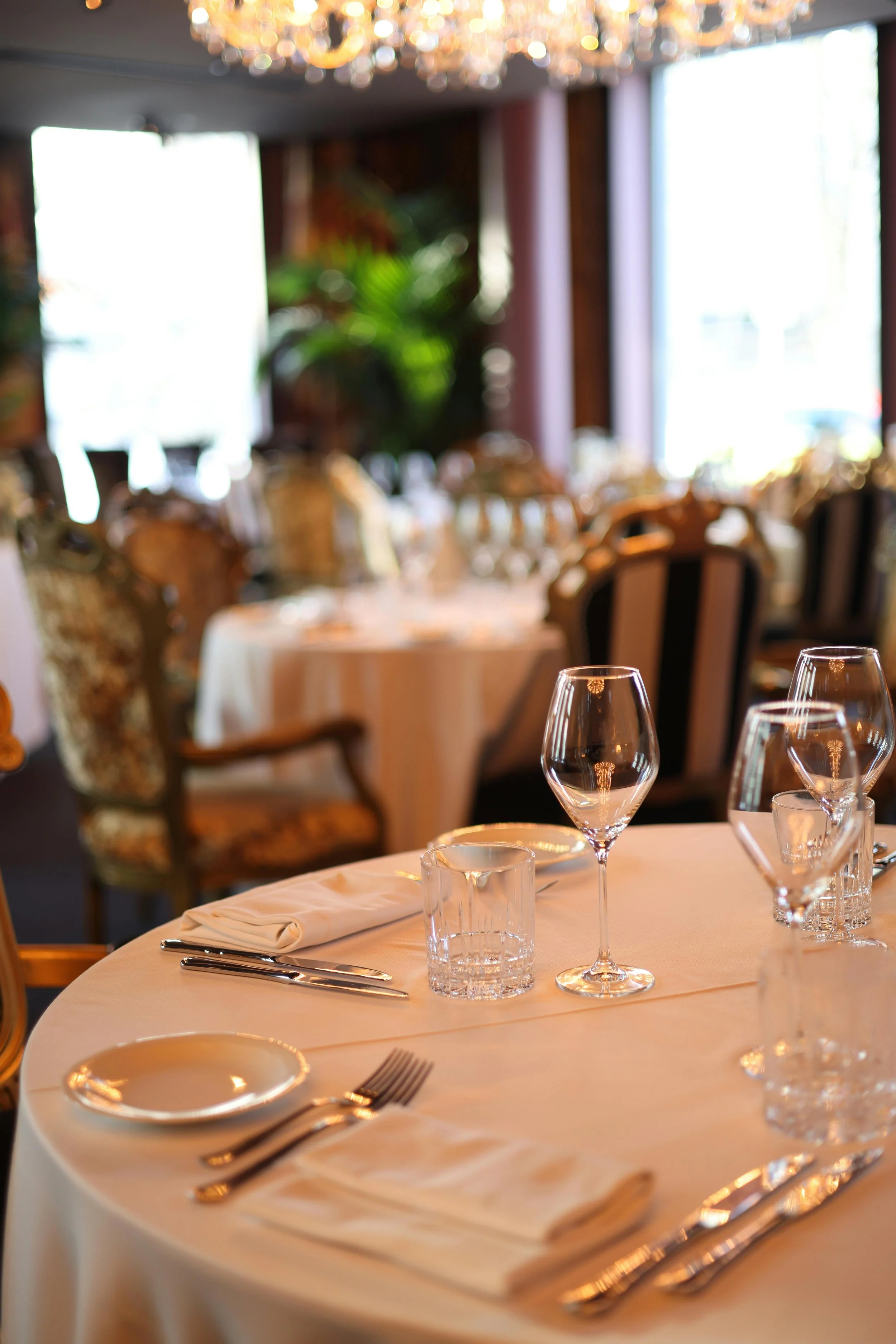 Elegant dining setup with white tablecloth, wine glasses, silverware, and a napkin, in a restaurant with large windows, wooden furniture, and chandelier lighting.