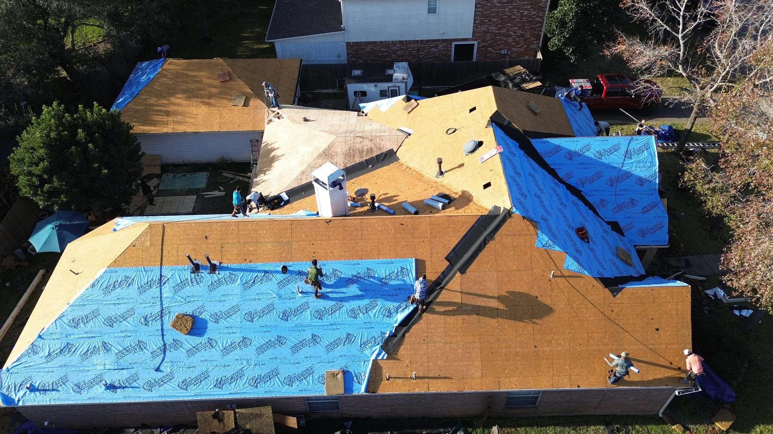 Aerial view of a house under construction with workers installing roofing materials. Part of the roof is covered with blue weatherproof underlayment, while other sections are still being worked on.