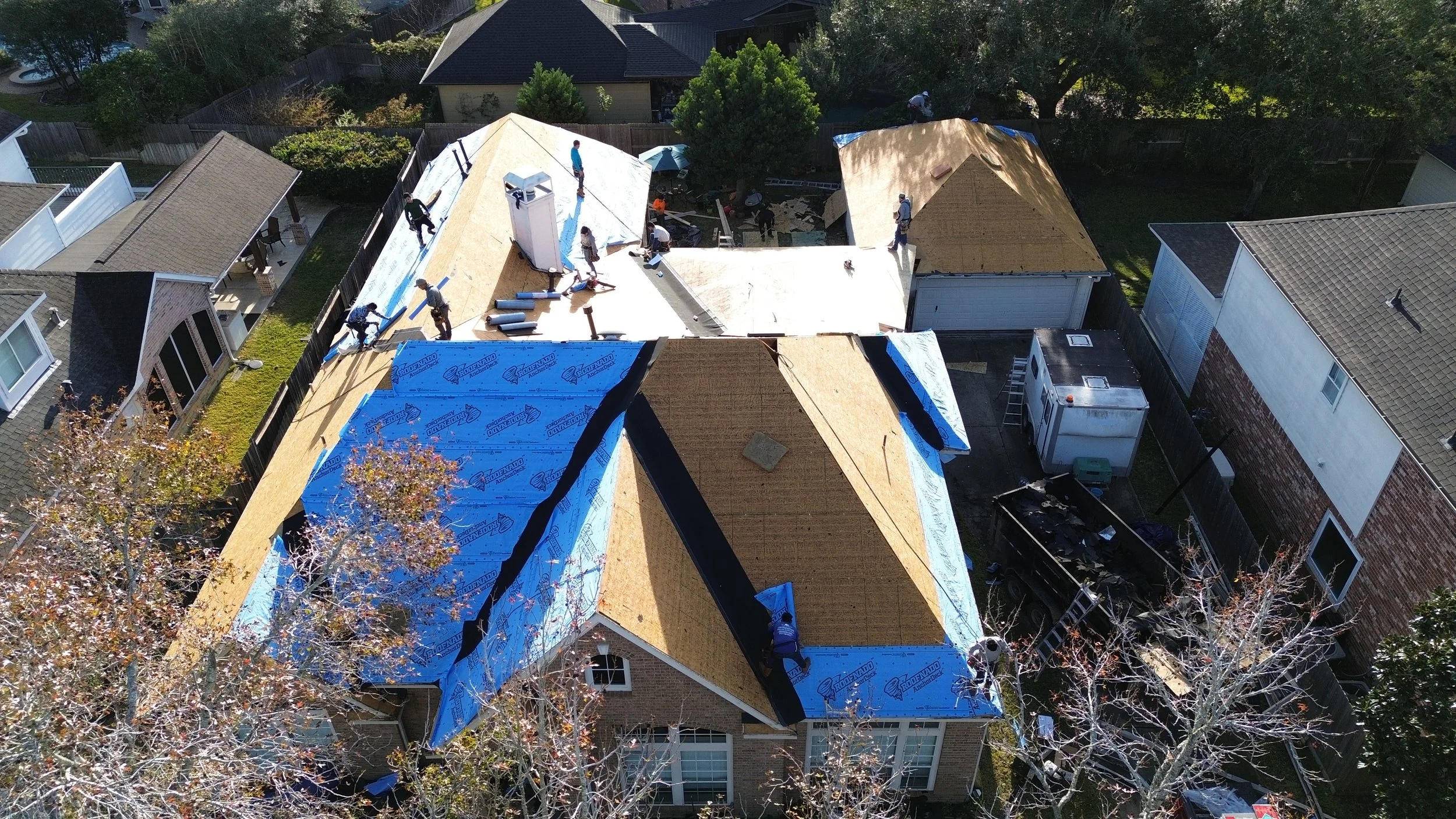 Aerial view of a house under construction with workers installing the roof, some with blue roofing material, in a neighborhood with trees, fences, and neighboring houses.