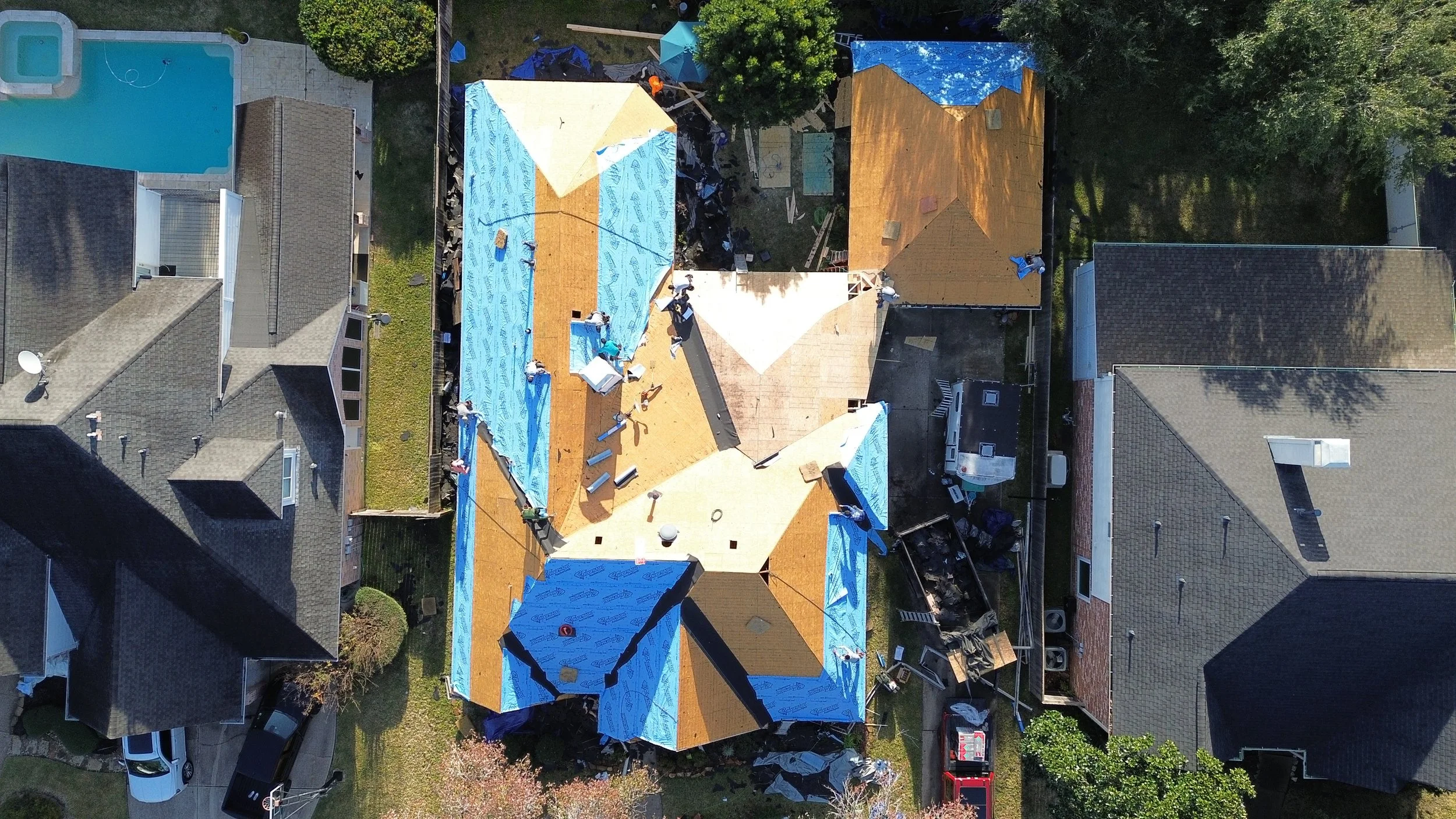 Aerial view of a house under construction with the roof partially covered in blue weatherproofing material, surrounded by neighboring houses with gray roofs, a swimming pool, and green trees.