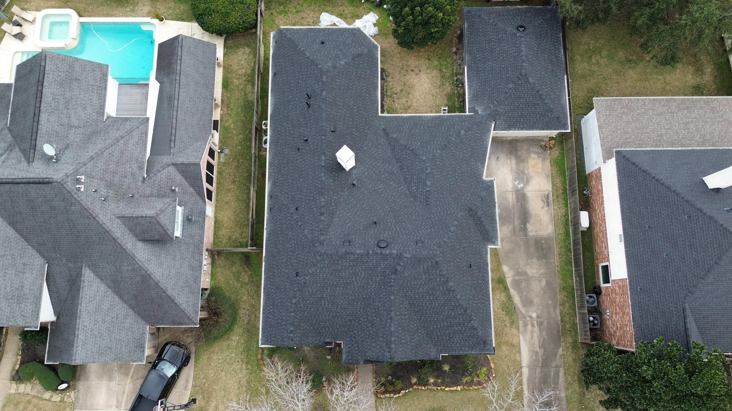 Aerial view of rooftops of several suburban houses, some with dark gray shingles, one with a small pool, yards, a driveway with a black vehicle, and trees.