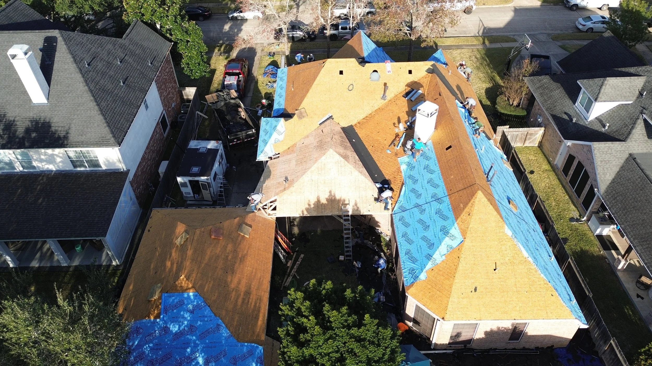 Aerial view of a residential construction site with workers installing roofing on a house, surrounded by neighboring houses and cars parked in driveways.