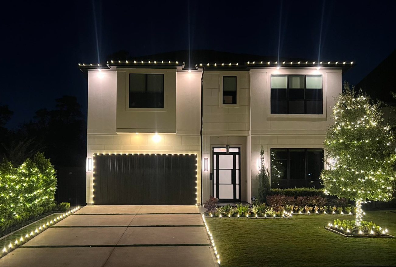 Modern two-story house decorated with Christmas lights at night, includes illuminated trees, bushes, and driveway.