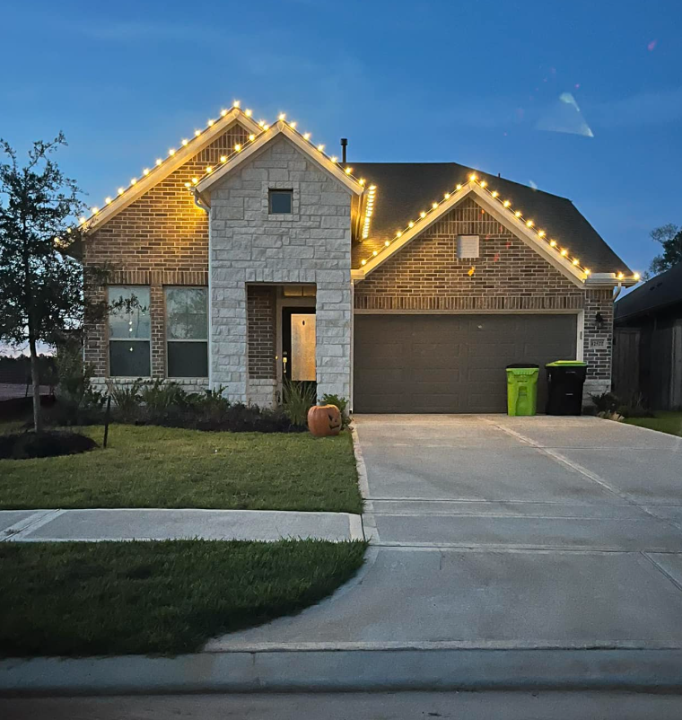 A two-story suburban house with brick and stone exterior, decorated with string lights along the roofline, and a small lawn with a pumpkin at the front. Two trash bins are near the garage on the driveway.