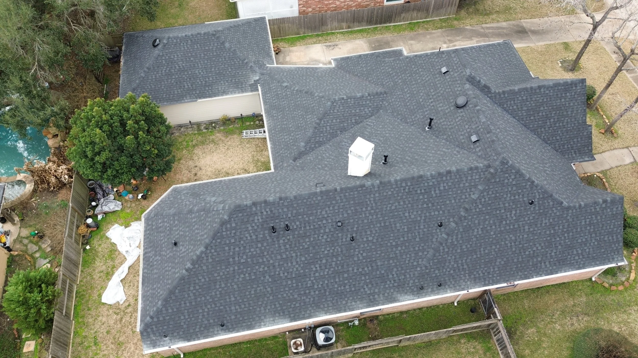 An aerial view of a residential house with a dark gray shingle roof, surrounded by a backyard with trees, a fence, and scattered outdoor items.