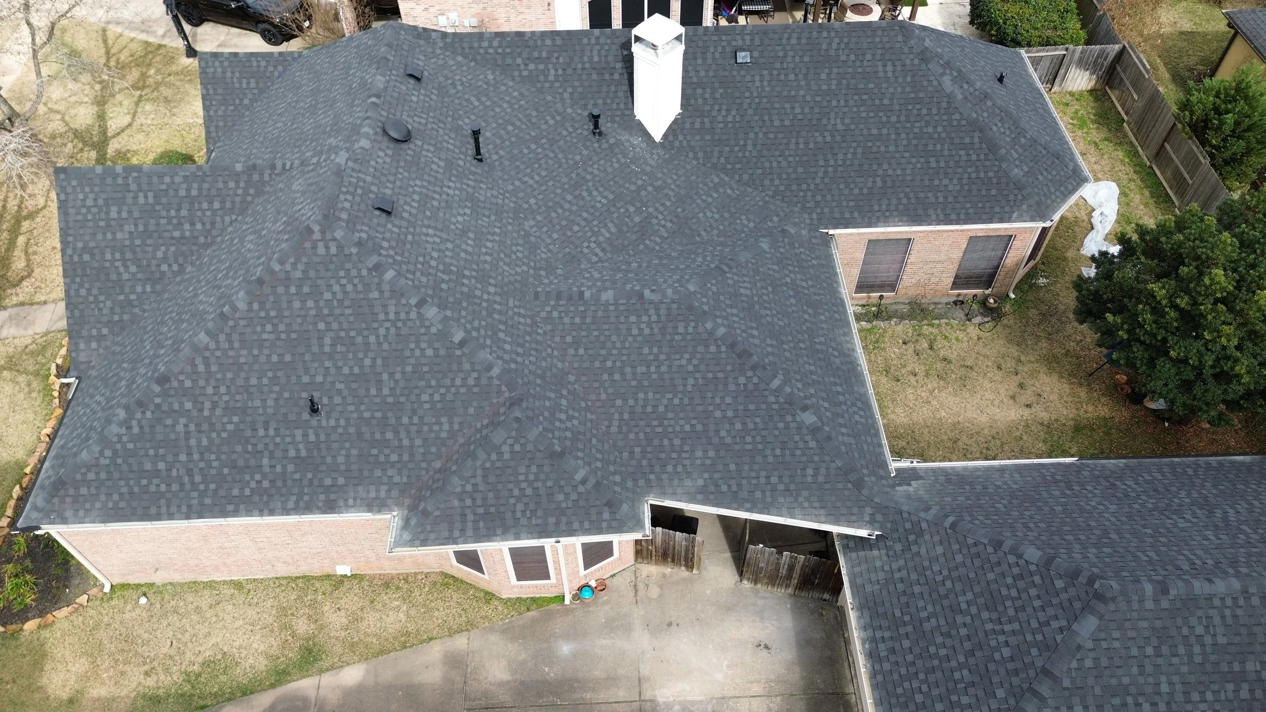 Aerial view of a house with gray asphalt shingle roof, chimney, and vents. Surrounding yard with grass, trees, and a driveway.