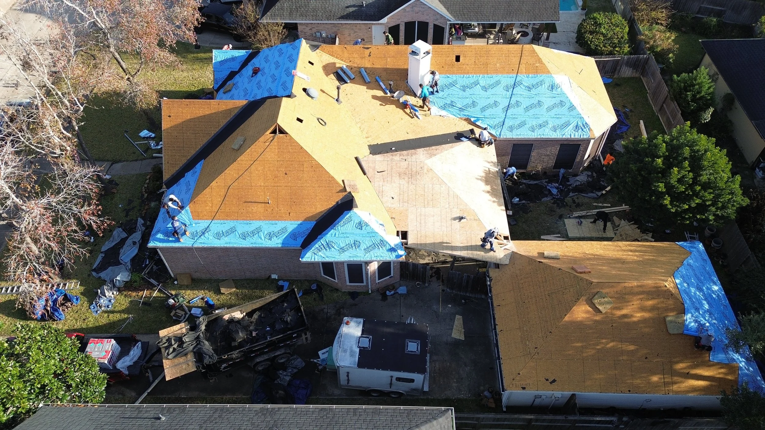 An aerial view of a house under construction, with workers installing a new roof that is partially covered with blue protective roofing material.