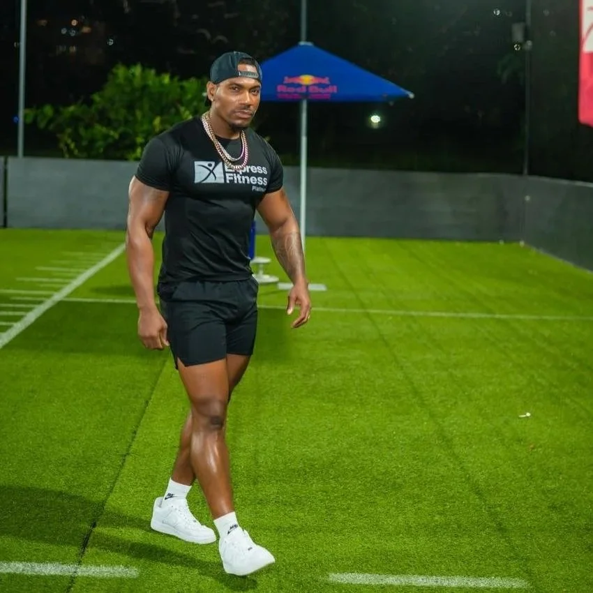 A man dressed in athletic gear is standing on a grassy sports field at night, wearing a cap backwards, a black workout shirt with a fitness logo, black shorts, white sneakers, and layered necklaces, with a Red Bull umbrella visible in the background.