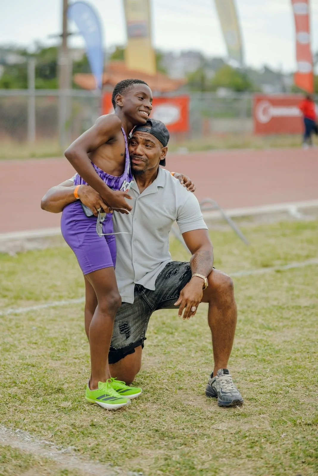 A young boy dressed in purple shorts and a tank top being lifted by a man in a white polo shirt and black shorts on an outdoor track field, both smiling and showing affection.
