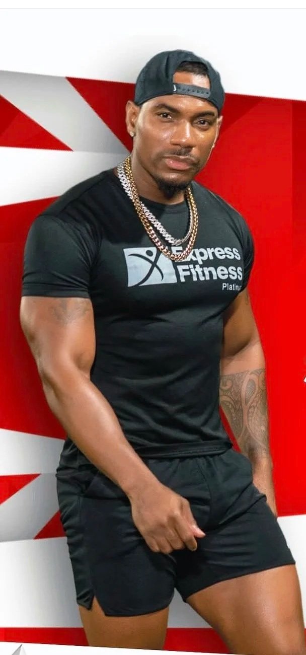 A muscular man wearing a black cap backwards, a black T-shirt with 'Xpress Fitness' logo, gold chains, and black shorts, standing against a red and white geometric background.