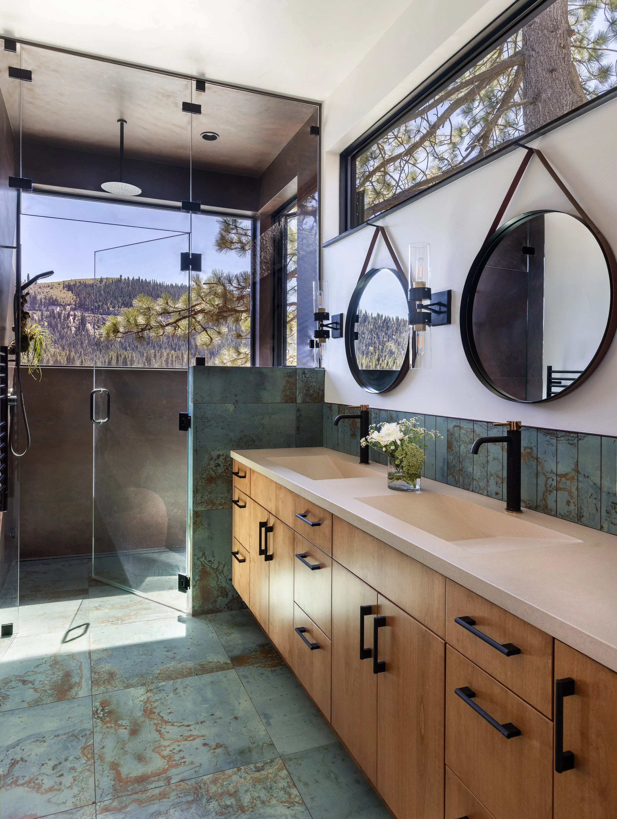 Modern bathroom with wooden vanity, beige countertop, black fixtures, large circular mirrors, and a walk-in shower with a glass door, mountain view outside, and natural light.