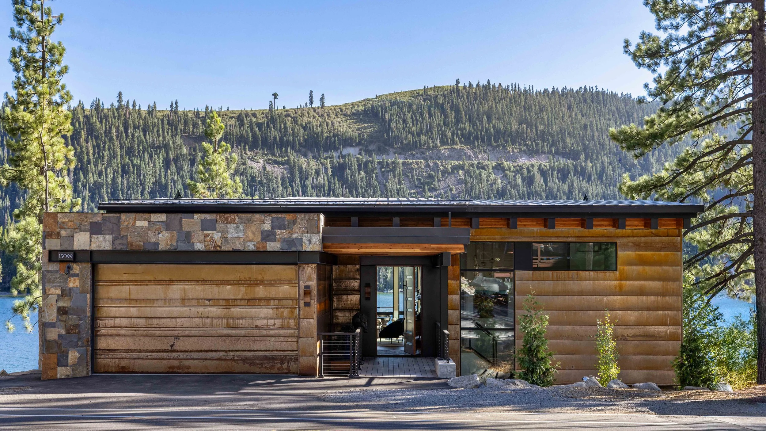 Modern house with stone and wood exterior in front of forested mountains and pine trees under a clear blue sky.