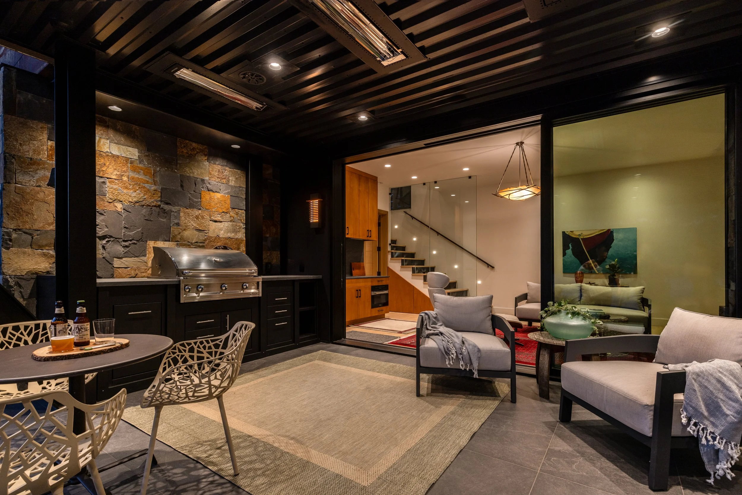 Interior of a modern open-plan living area featuring a kitchen with stone wall, wooden cabinets, and a staircase, adjacent to a cozy seating space with sofas, chairs, and a dining table, decorated with a chandelier and artwork.