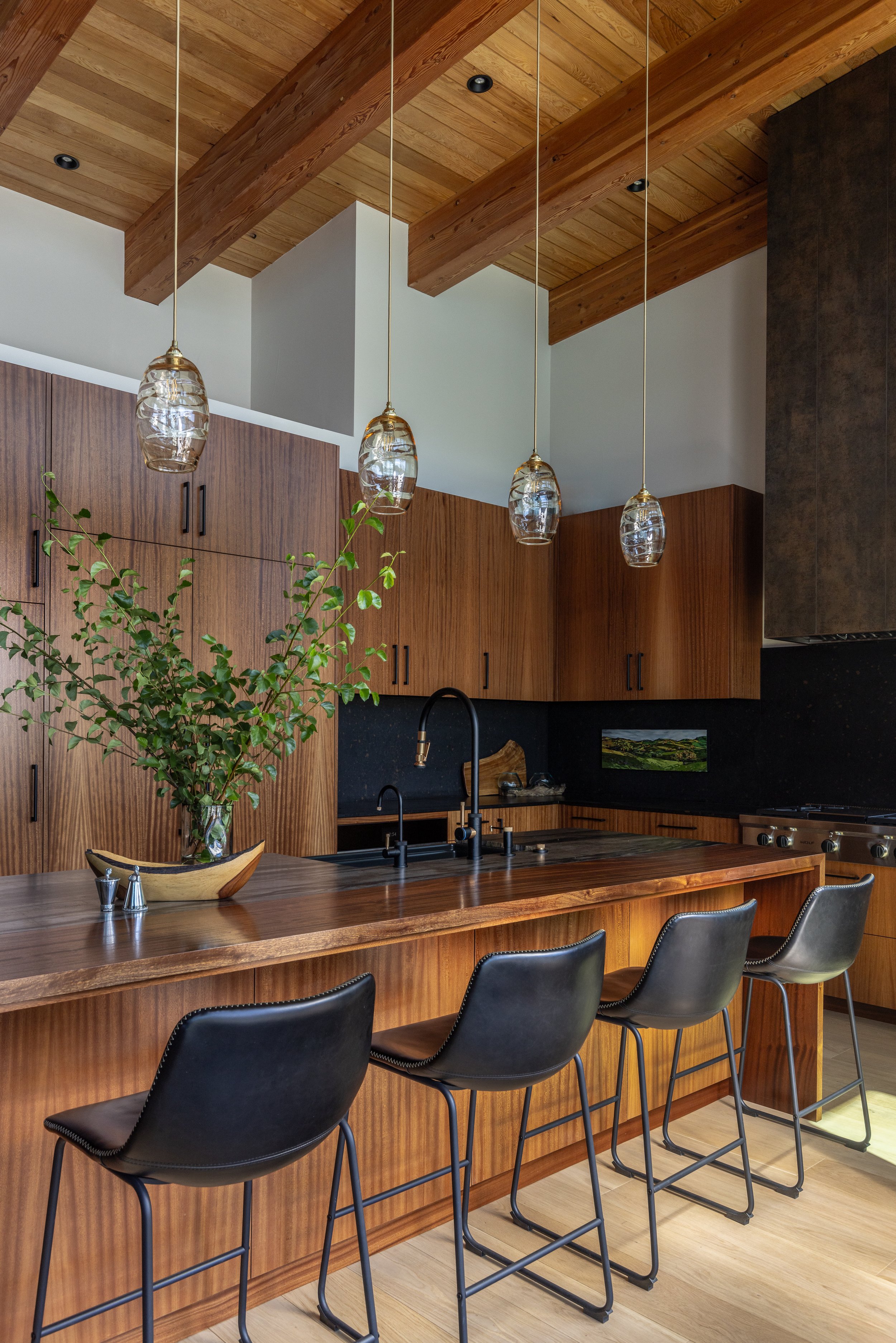 Modern kitchen with wooden cabinets, hanging pendant lights, a breakfast bar with black chairs, and a decorative plant on the counter.