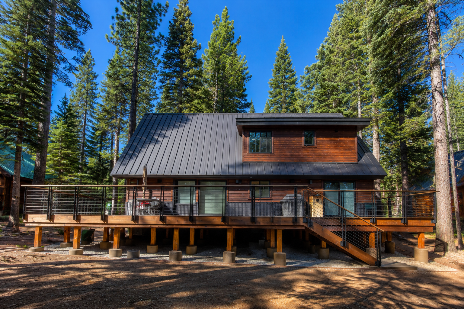 Wooden house on stilts with a metal roof, surrounded by tall pine trees, in a forested area under a clear blue sky.