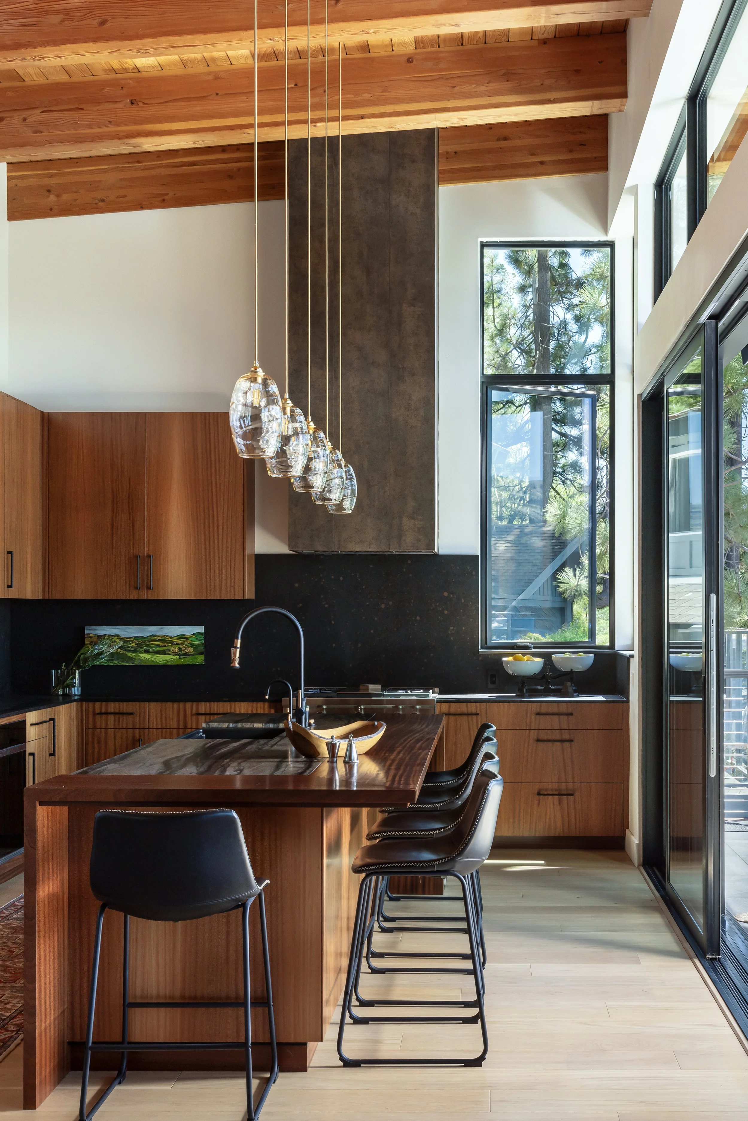 Modern kitchen with wooden cabinetry, pendant lights, black countertops, and large windows showing trees outside.
