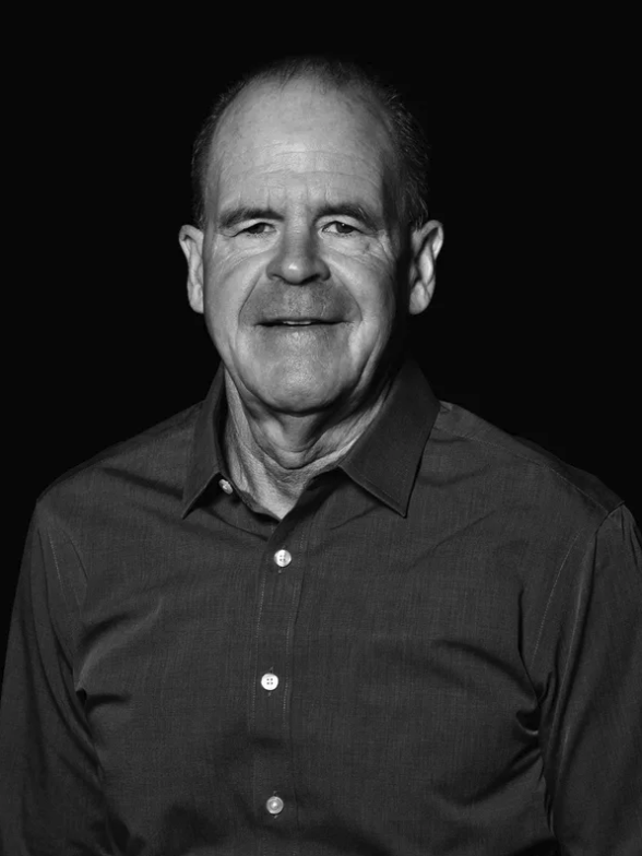 Black and white portrait of an older man with short hair, wearing a button-up shirt, smiling slightly, against a plain dark background.