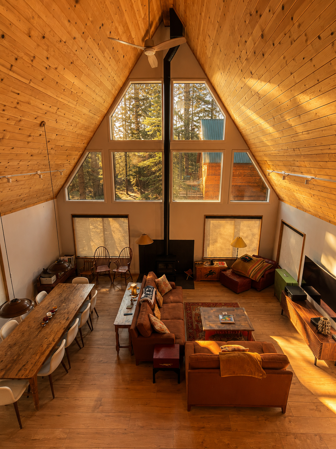 A spacious living room with high, angled wooden ceiling, large triangular windows showing trees outside, a wood stove, a sofa with throw pillows, a coffee table, two chairs, and a dining table with chairs.