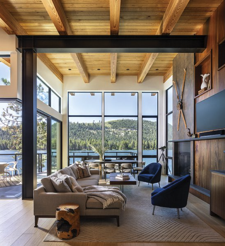Living room with floor-to-ceiling windows showing a lake and forest, wooden ceiling, modern furniture, and wall decor including crossed oars.