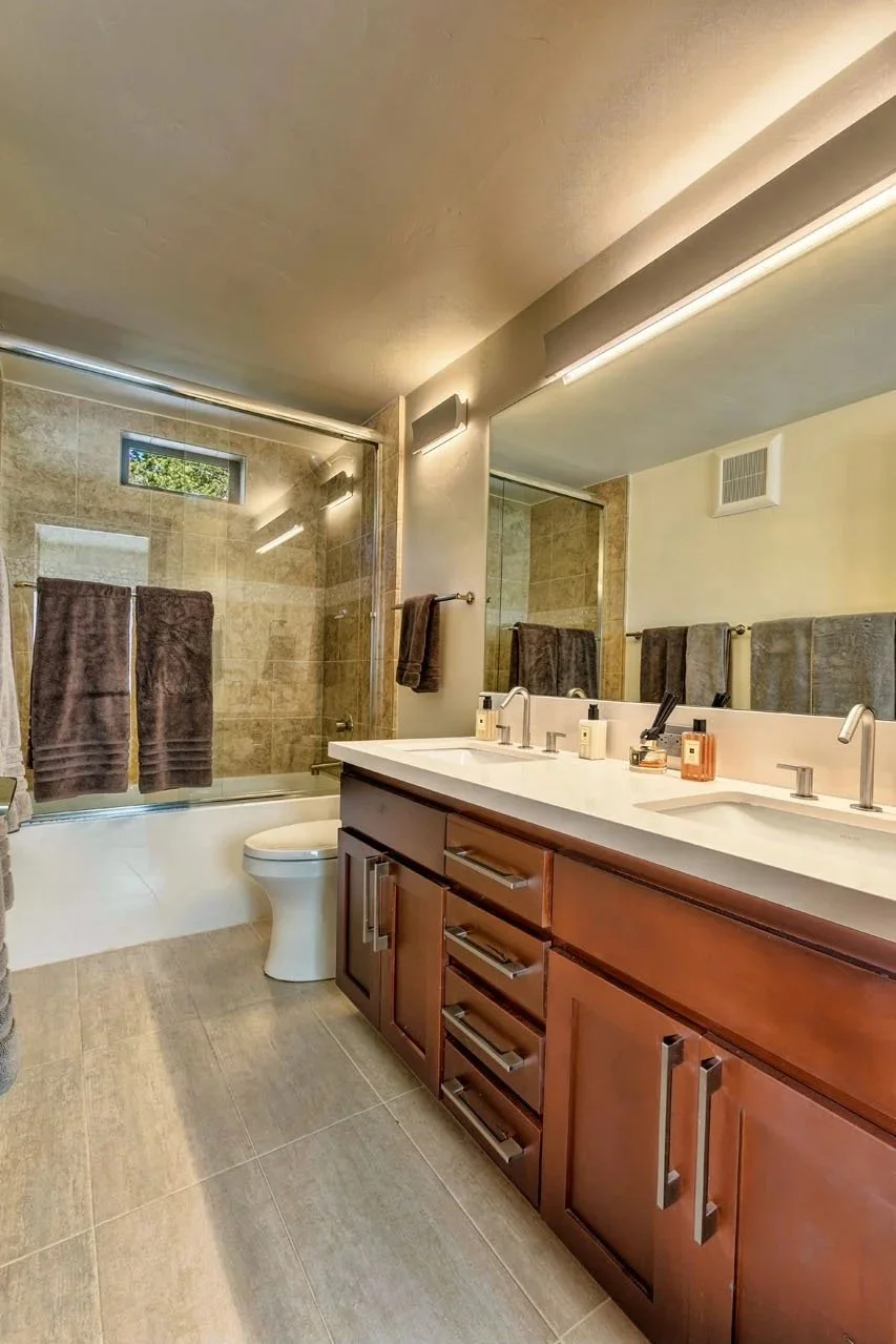 Modern bathroom with dual sinks, wooden cabinet, large mirror, and a glass-enclosed shower area with brown towels.