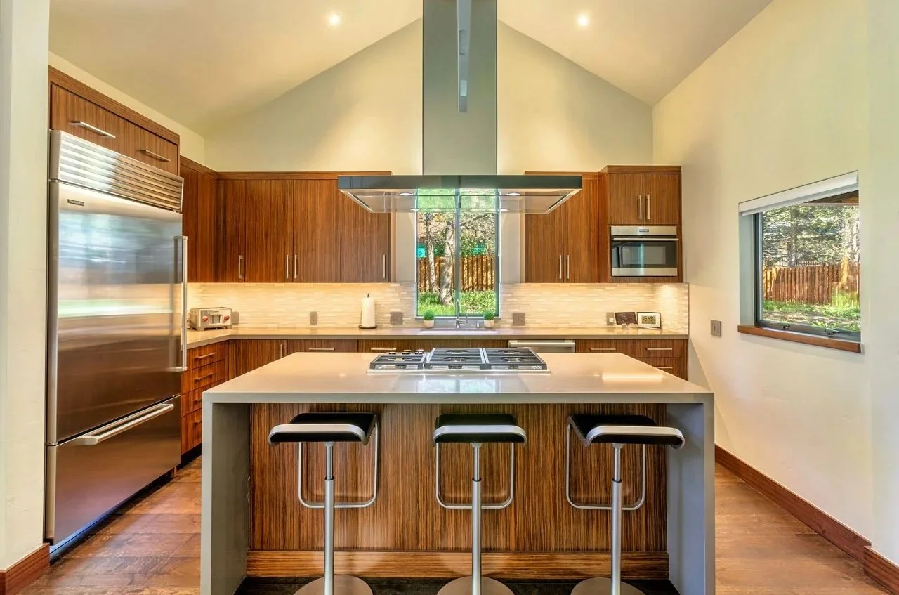 Modern kitchen with wooden cabinets, a large island with a gas stove, stainless steel appliances, and a window showing a backyard with trees and a wooden fence.