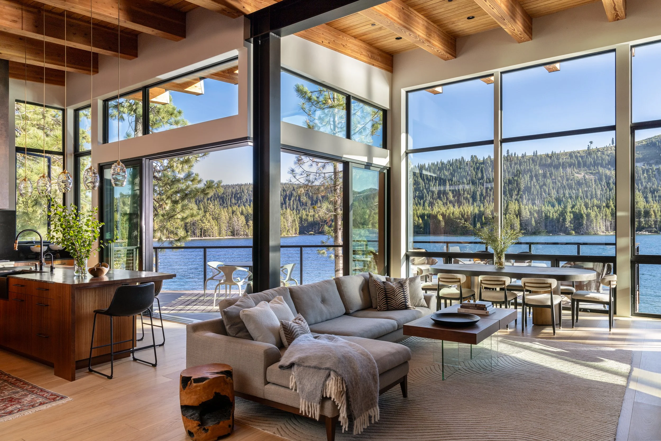 Modern living room with large floor-to-ceiling windows overlooking a lake and forested hills, featuring a gray sectional sofa, wooden coffee table, and decorative lighting fixtures.