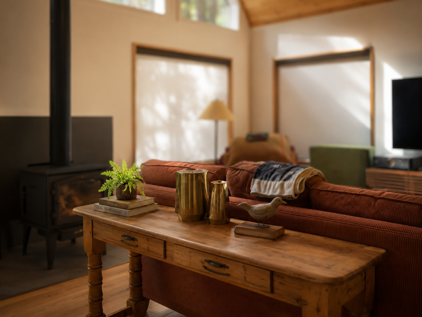 Living room with a wooden coffee table, a cozy red sofa, decorative gold vases, a potted fern, a bird figurine, a wood stove, a TV, and large windows with sheer curtains letting in natural light.