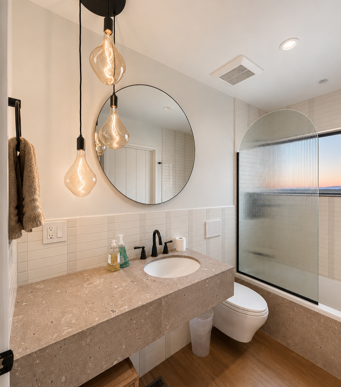 Modern bathroom with a beige marble vanity, black fixtures, a round mirror, hanging pendant lights, a frosted glass shower door, a window with a sunset view, and beige tiled walls.