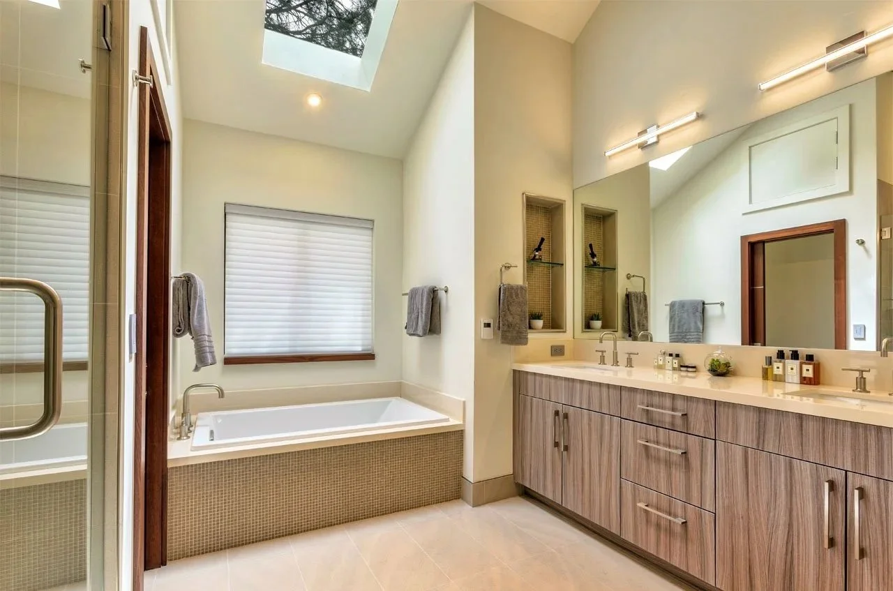 Bright modern bathroom with a bathtub, large mirror, dual sinks, gray towels, small decorative items, window with closed blinds, and a skylight in the ceiling.