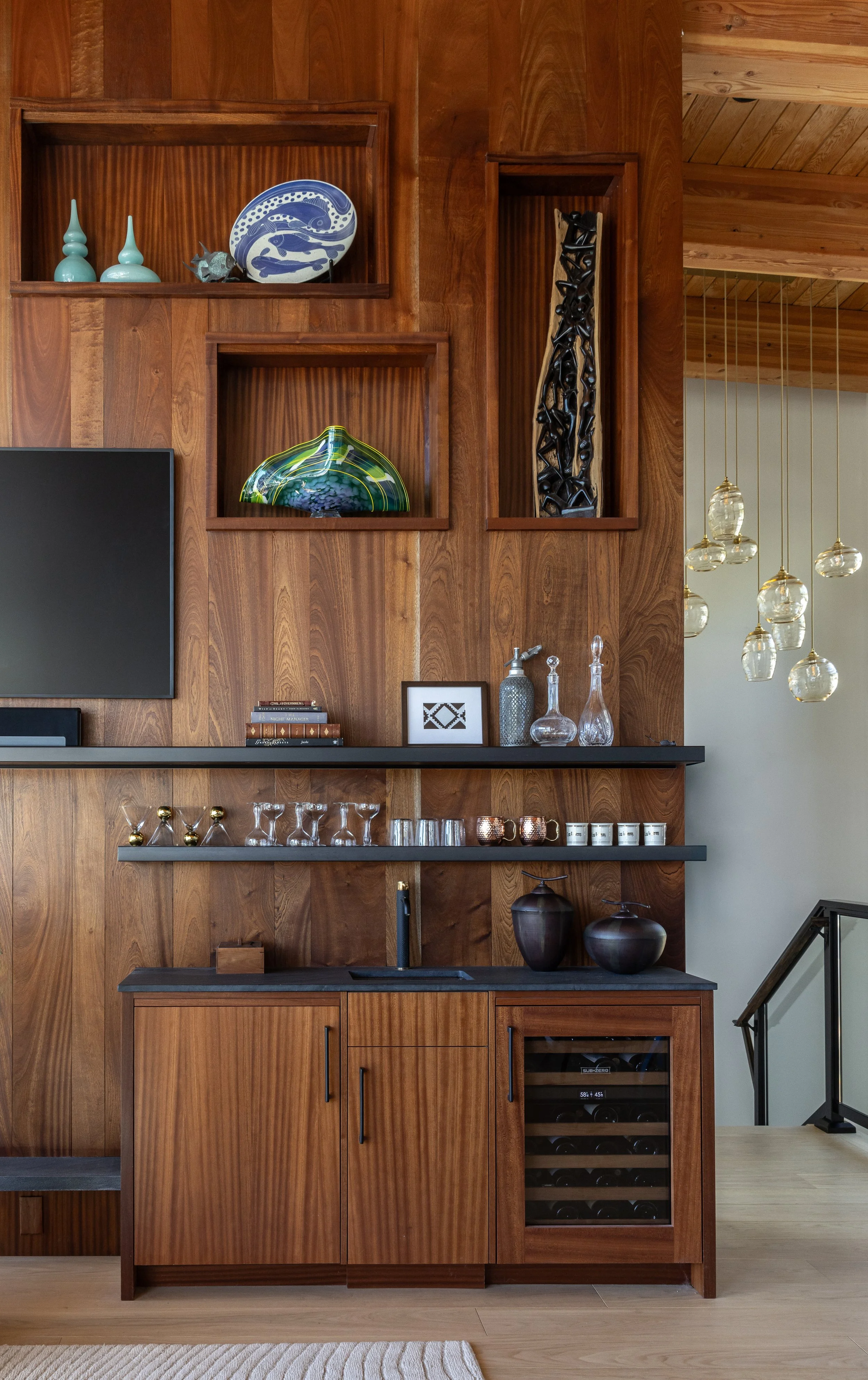 A wooden wall with built-in shelves displaying decorative items such as vases, a plate with fish design, and art pieces. Below is a black countertop with glassware and decorative objects. To the right, there is a wine fridge with bottles inside, and a staircase with a black railing is visible in the corner. Hanging light fixtures with glass globes are also seen in the background.