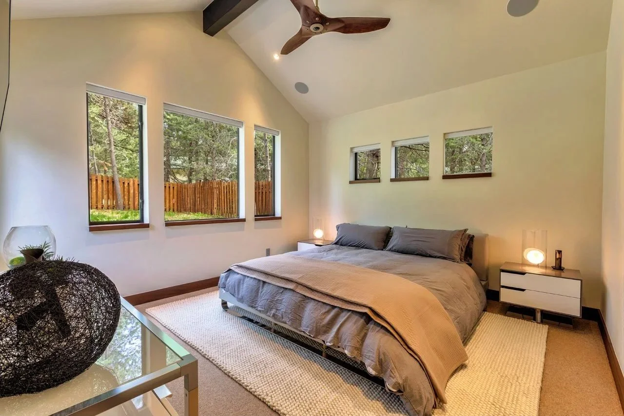 A bedroom with a sloped ceiling, three large windows showing a view of trees and a wooden fence, and four small windows above the bed. The bed has gray bedding and a beige blanket, with two nightstands on either side, each with a small lamp. There is a ceiling fan with wooden blades and a white textured rug on the beige carpet floor.