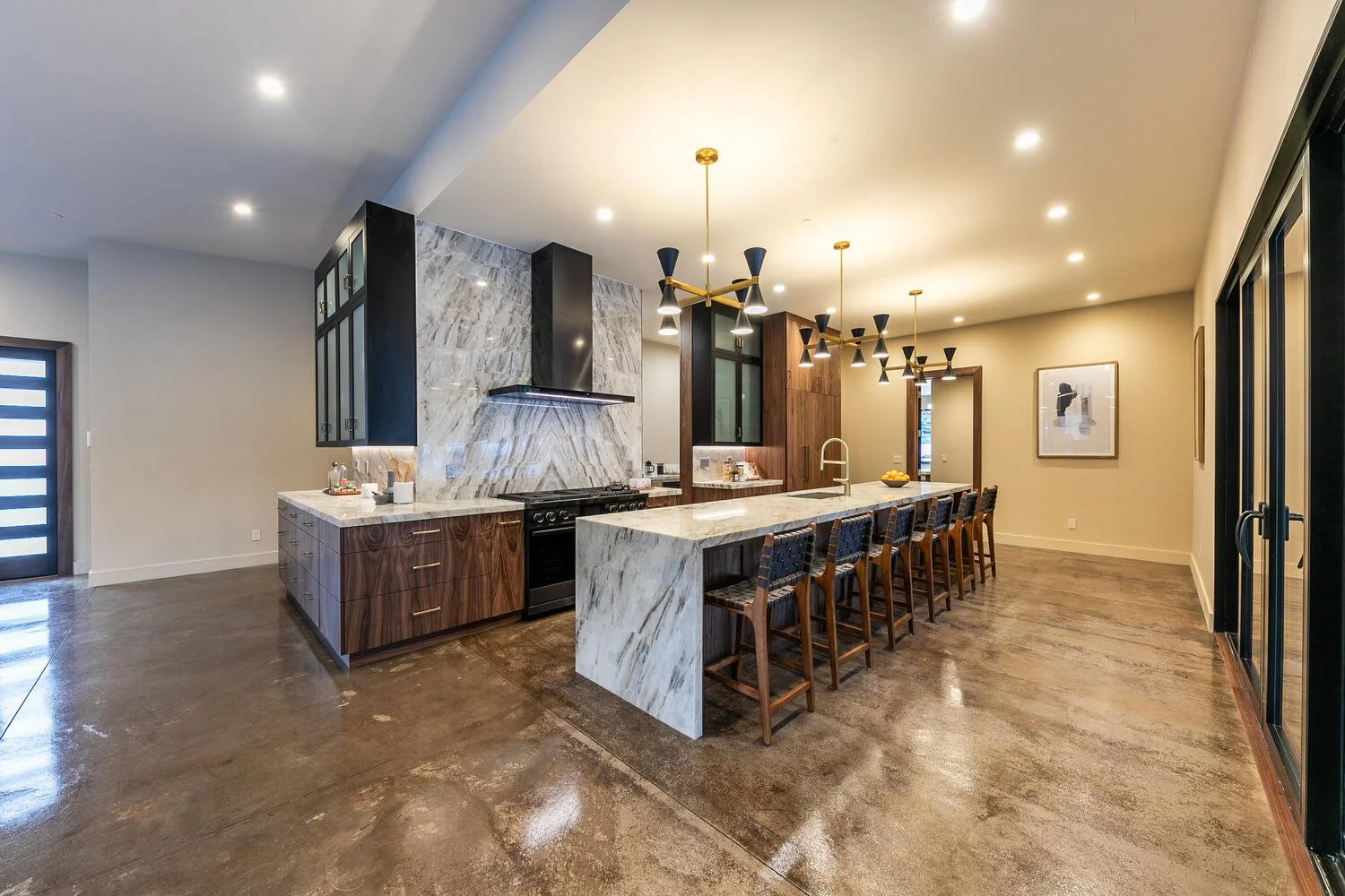 Modern kitchen with a large marble island, wooden cabinetry, a black and gold chandelier, and a polished concrete floor.