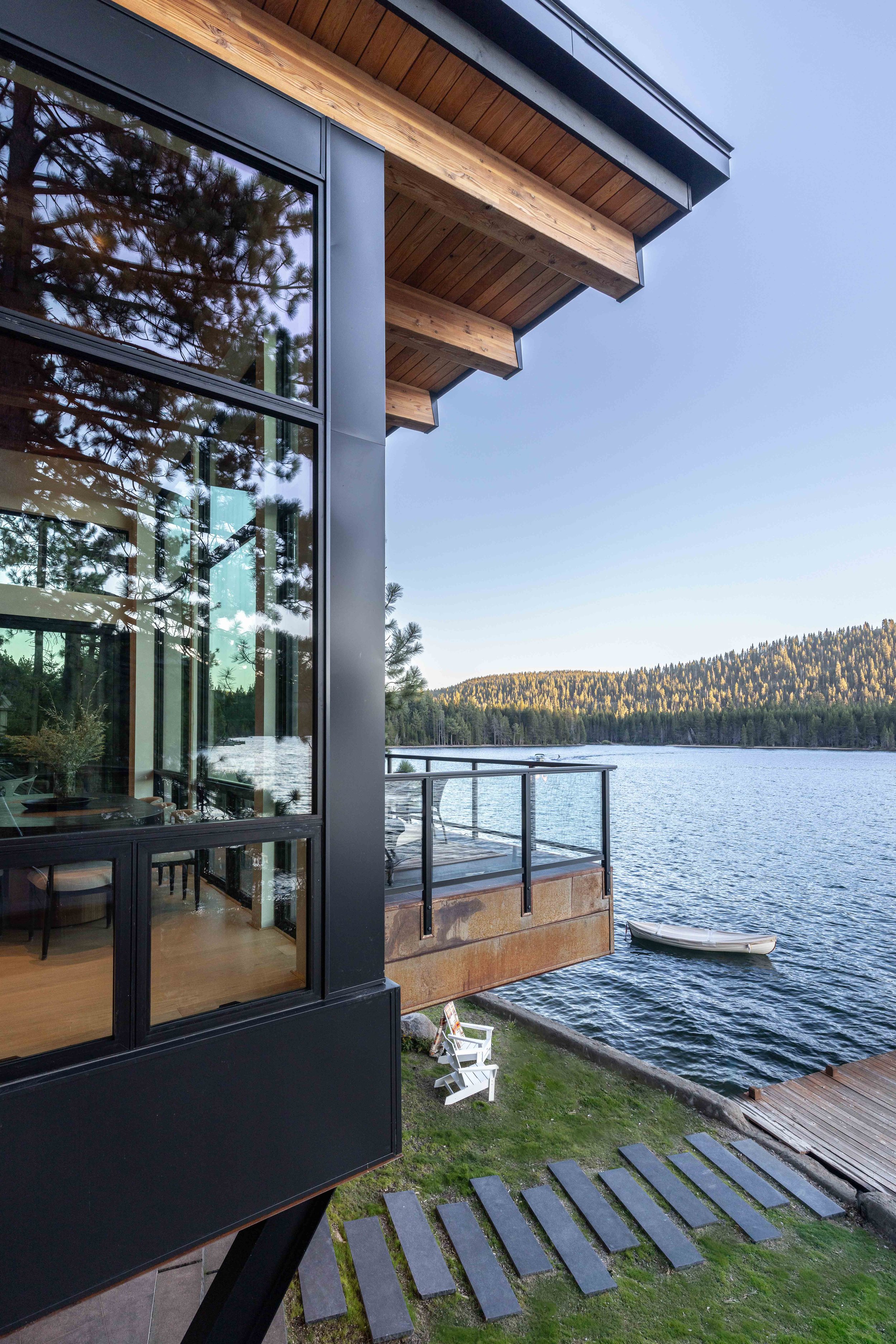 Modern building with large glass windows overlooking a lake, with trees on the opposite shore and outdoor furniture including chairs, a small boat at the water's edge, and a scenic mountain background under a clear sky.