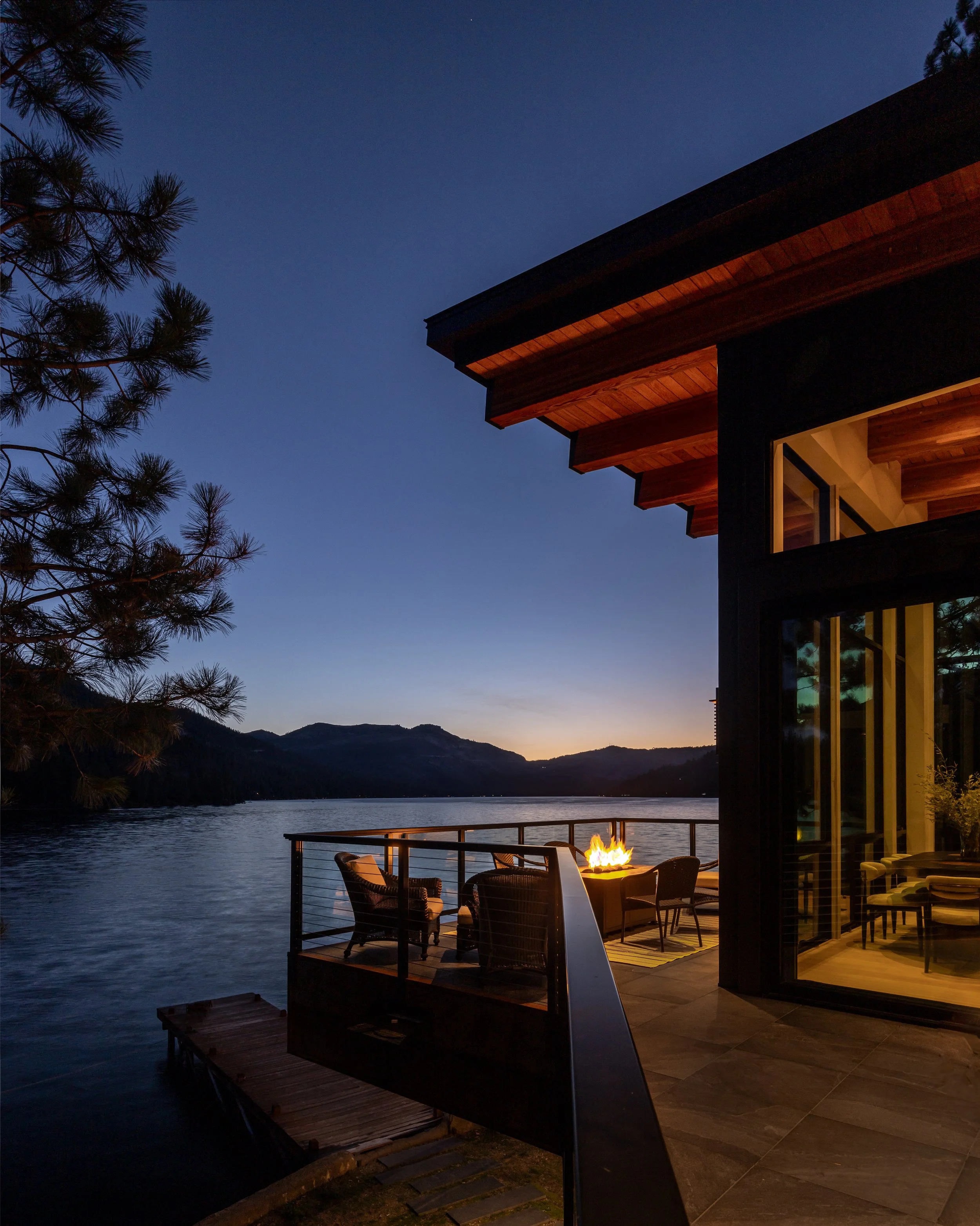 A lakeside patio at dusk with a firepit and outdoor seating, overlooking a body of water with mountain silhouettes in the background. House with large windows and a wooden roof is partially visible on the right.