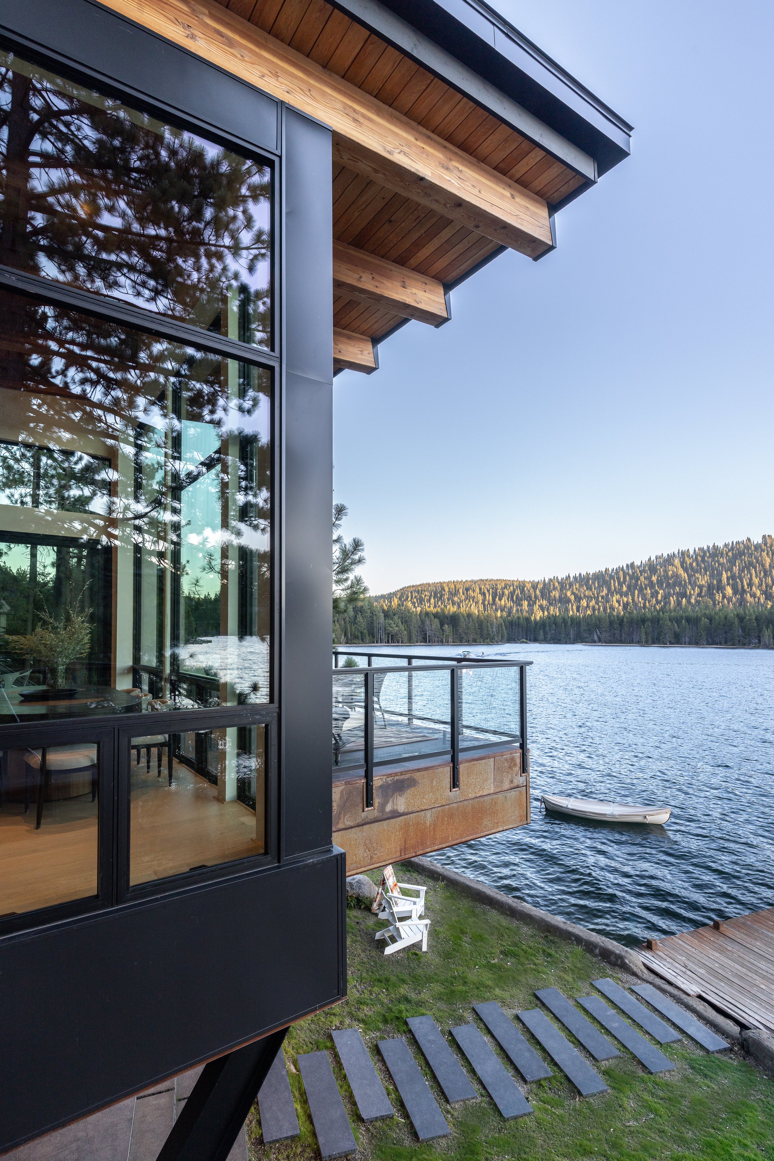 A modern building with large windows and wooden overhangs situated by a lake. The view includes a small boat on the water, a grassy area with a white chair, and forested hills in the background.