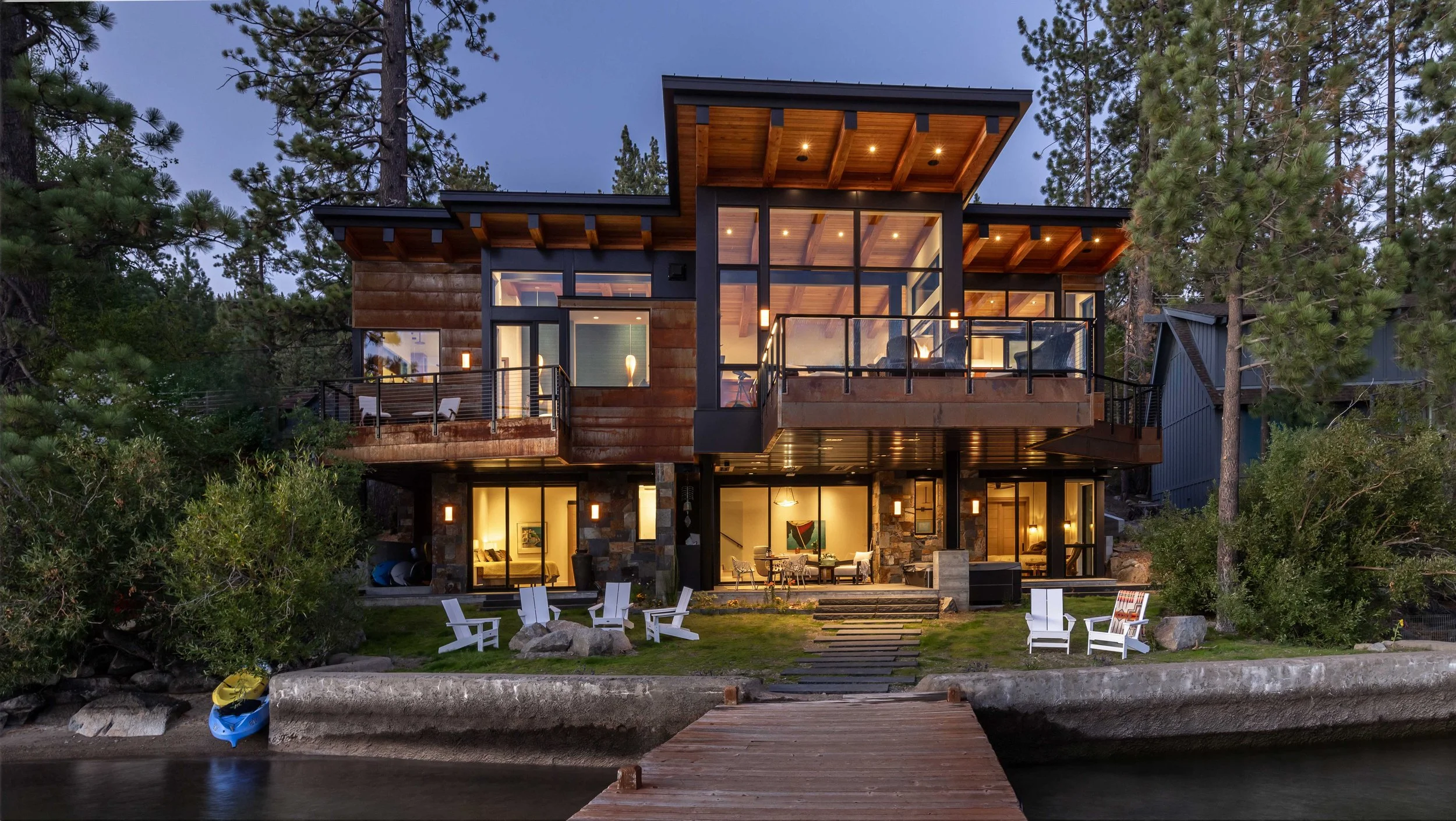 Modern multi-story house with large glass windows and wooden accents, situated among trees near a body of water at dusk, with outdoor seating and a dock in the foreground.