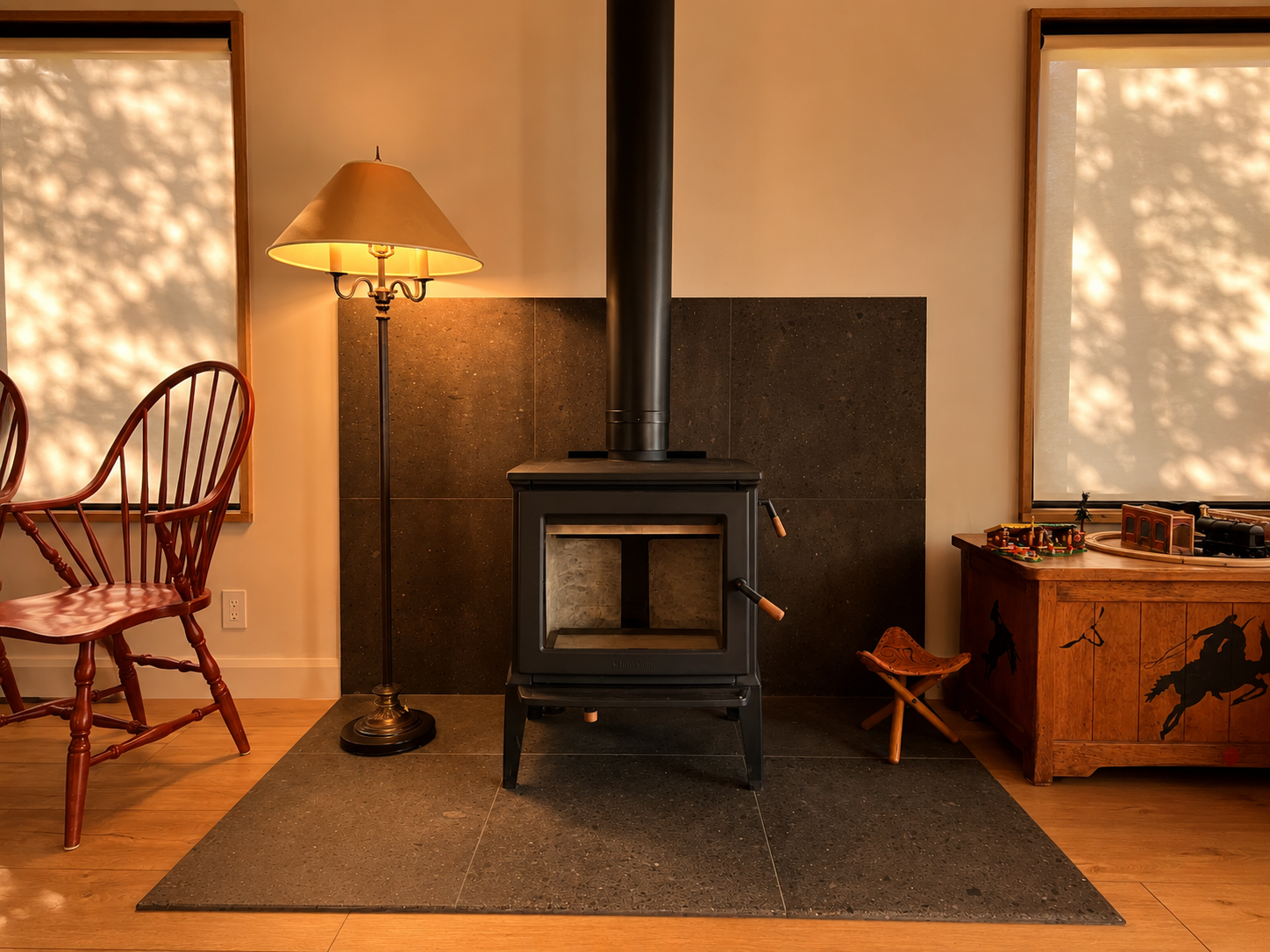Living room with a wood stove centered on a dark tile hearth, flanked by a tall floor lamp on the left and a wooden playtable with toy trains on the right, with chairs and large windows on either side.