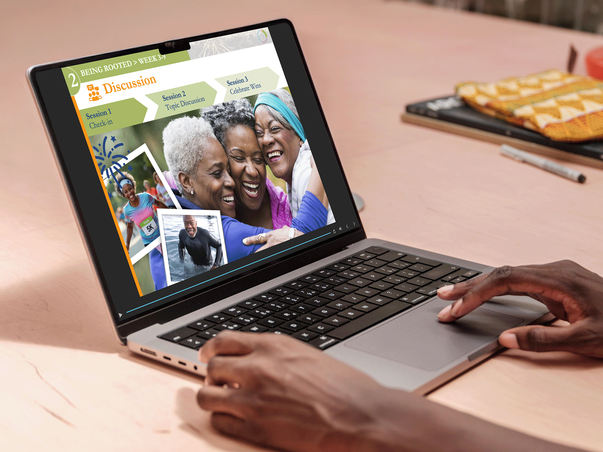 A person's hand is using a laptop displaying a presentation slide about a discussion group, featuring photos of older women and a young girl, on a wooden desk with a notebook and pen nearby.