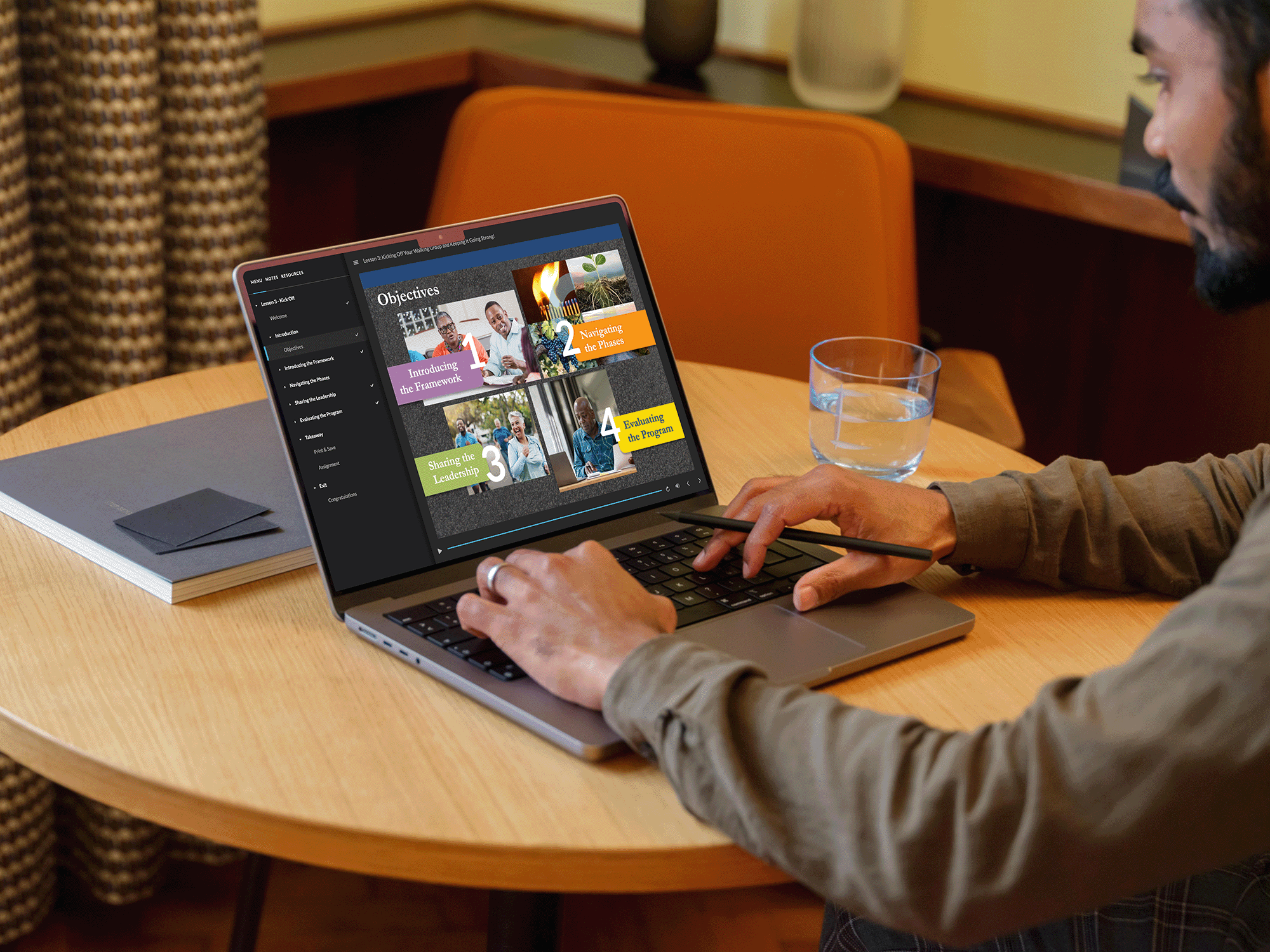 A man using a stylus to interact with a laptop displaying an online course with colorful objectives slides on a wooden table. There is a glass of water and a notebook nearby.