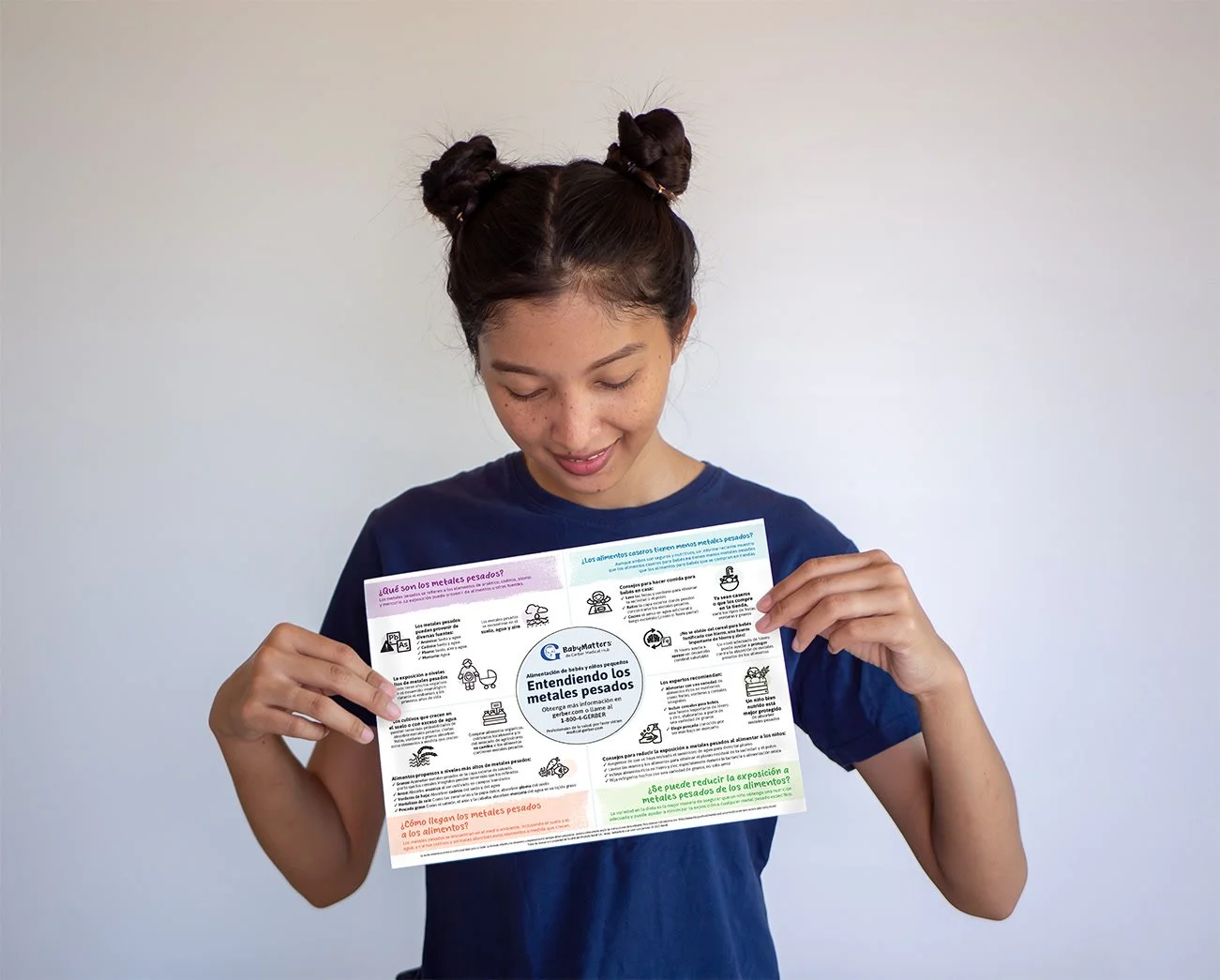 A young girl with two space buns looks down at a colorful informational flyer about heavy metals in foods. She is wearing a navy blue t-shirt and stands against a plain white wall.