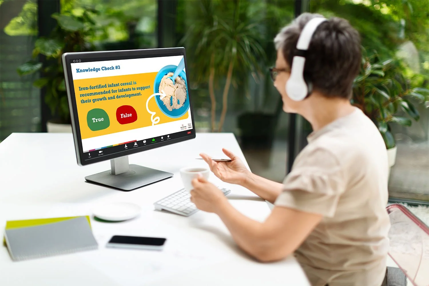 Woman participating in a virtual quiz or quiz-style lesson on a computer, with a question about infant cereal displayed on the screen, in a well-lit room with plants in the background.