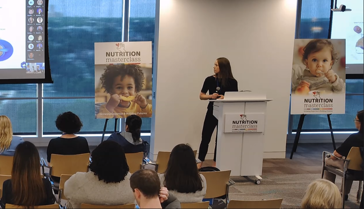 A woman stands at a podium giving a presentation to an audience about nutrition, with two large posters of children eating behind her, and a large screen displaying a virtual meeting on the left side.
