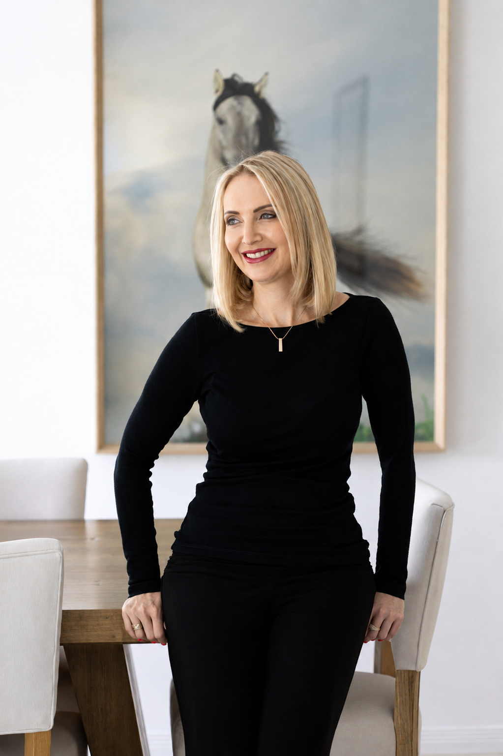 A woman with blonde hair wearing a black long-sleeve top and black pants, smiling, standing in a room with a large framed painting of a horse in the background.