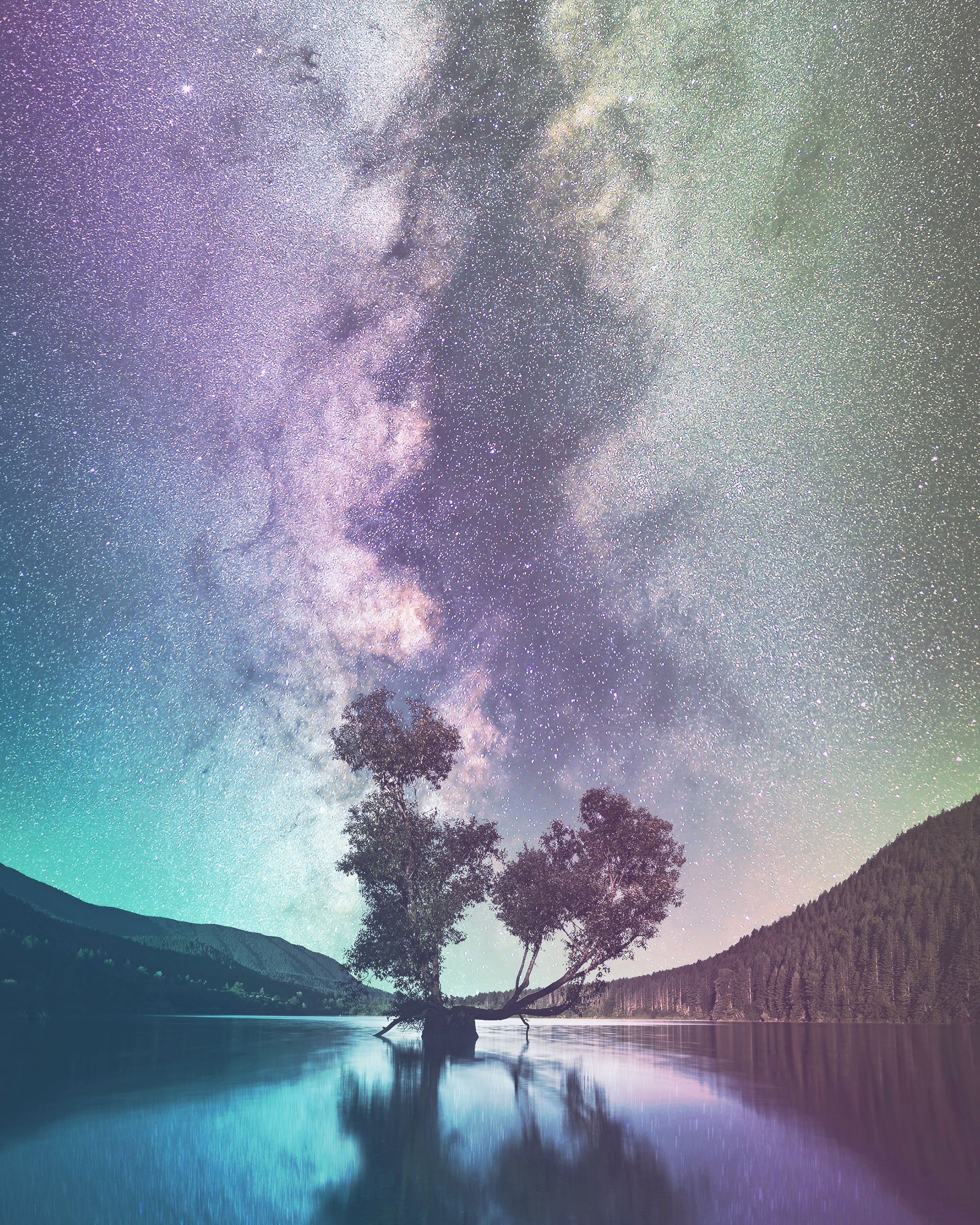 Night sky filled with stars and the Milky Way galaxy above a lake with trees and mountains along the shoreline, reflecting the starry sky.
