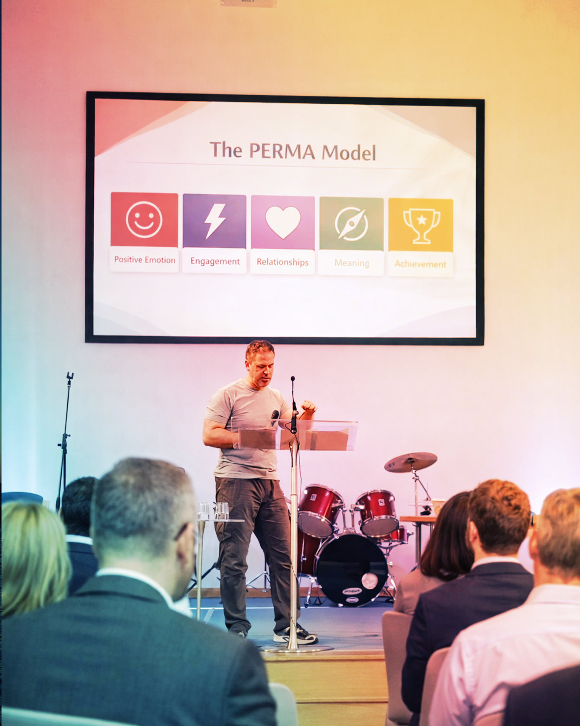 Ewan Innes giving a presentation on a stage with a large screen behind him displaying the title 'The PERMA Model' and icons representing Positive Emotion, Engagement, Relationships, Meaning, and Achievement. The audience is seated and listening.