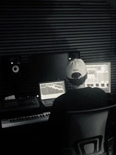 Person sitting at a music production workstation with multiple monitors and audio equipment, wearing a baseball cap, in a dark room with soundproofing panels.