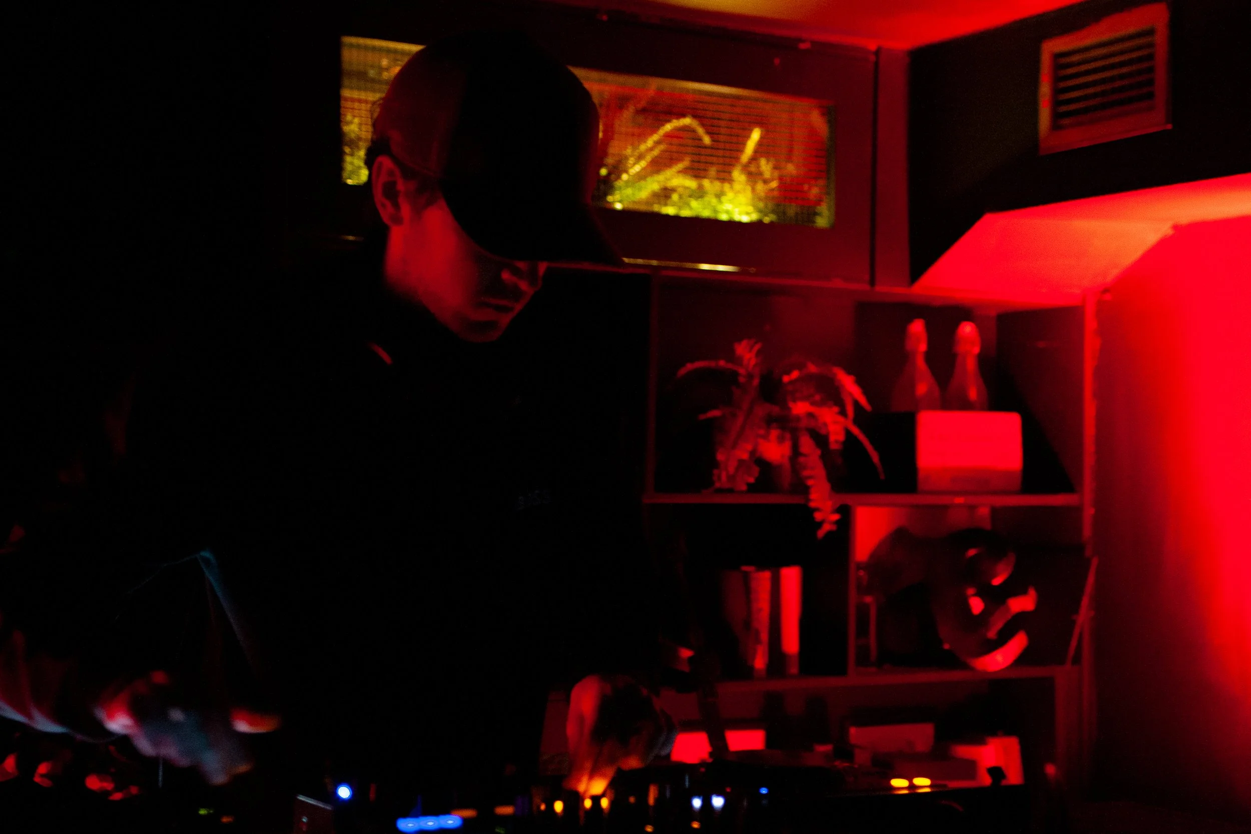 A person with glasses and a cap is seen in a dimly lit room with red lighting, working at a DJ deck with colorful controls.