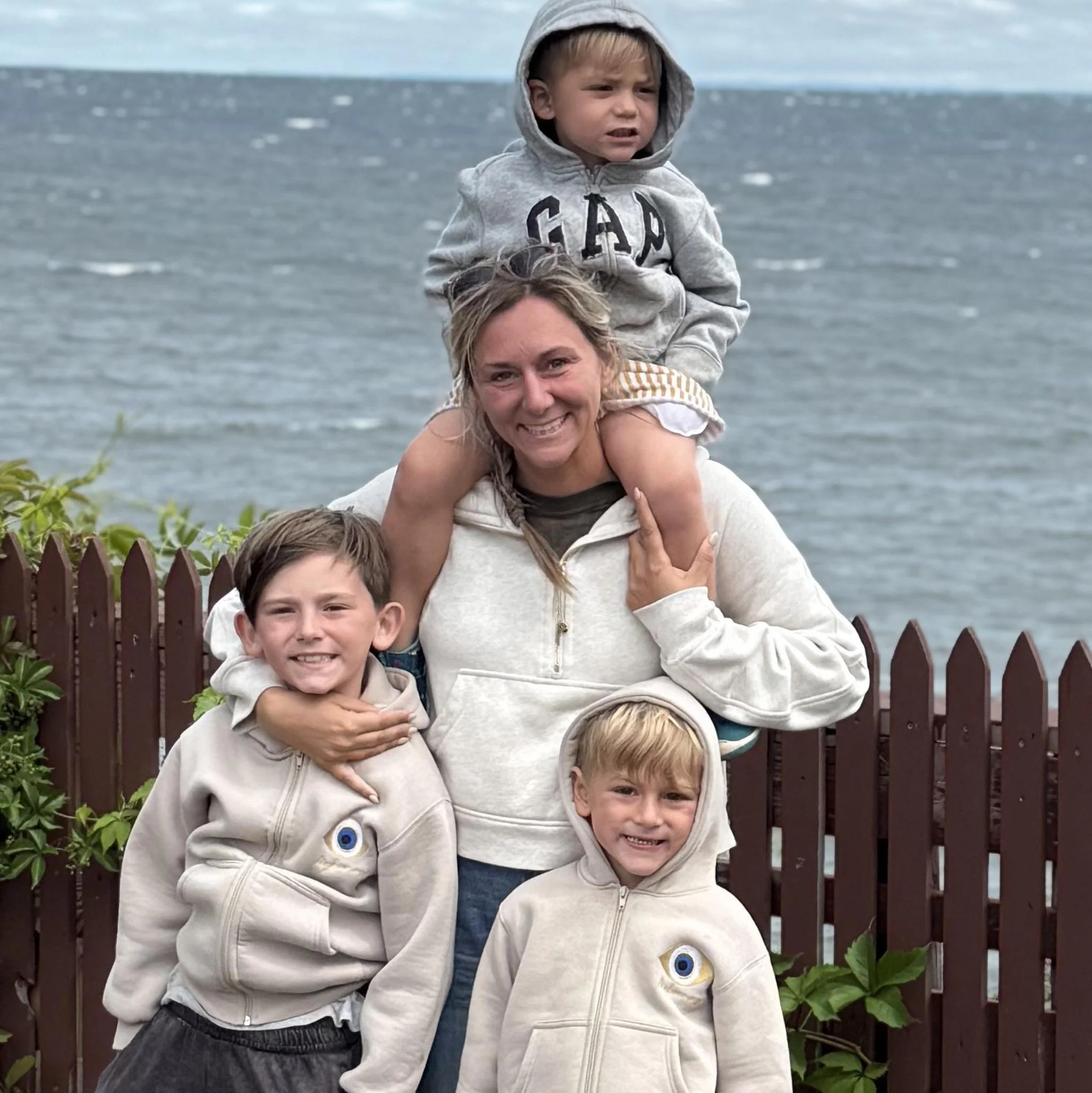 A woman with three children at the beach, one girl sitting on her shoulders wearing a gray hoodie with 'GAP' logo, and another girl and boy standing in front, all smiling. A wooden fence and green plants are in the background with a body of water behind.