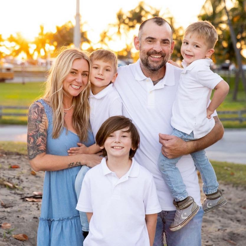 A smiling family of five outdoors during sunset, with a woman wearing a blue dress, a man in a white shirt holding two young boys in white shirts, and a young girl in a white shirt, all standing on a park trail with trees and a wooden fence in the background.