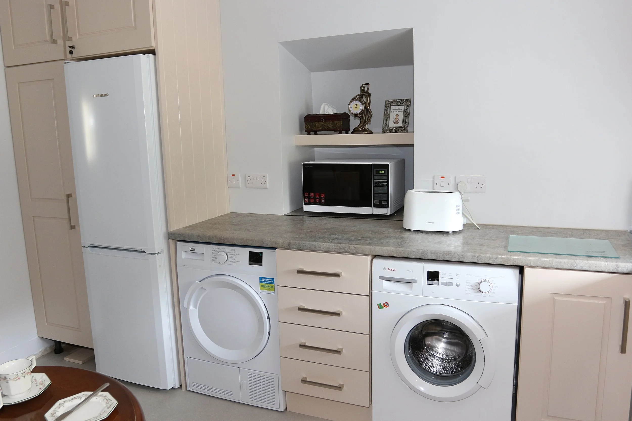 A kitchen with various appliances on a beige countertop, including a microwave oven and a toaster. Below the counter are a washing machine and a dryer. To the left, there is a tall white refrigerator. Above the microwave, a small decorative shelf holds a clock, a tissue box, and a framed sign. The background shows a plain white wall and beige cabinetry.